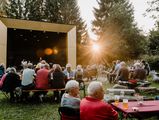 Publikum sitzt vor der Bühne des Musikpavillons im Kurpark Schierke und verfolgt die Veranstaltung zum Schierker Musiksommer. Der Pavillon erstrahlt im Gegenlicht der Sonne.